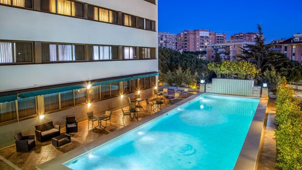 Una piscina con sillas de descanso y mesas, rodeada por un edificio de varios pisos y ventanas.
