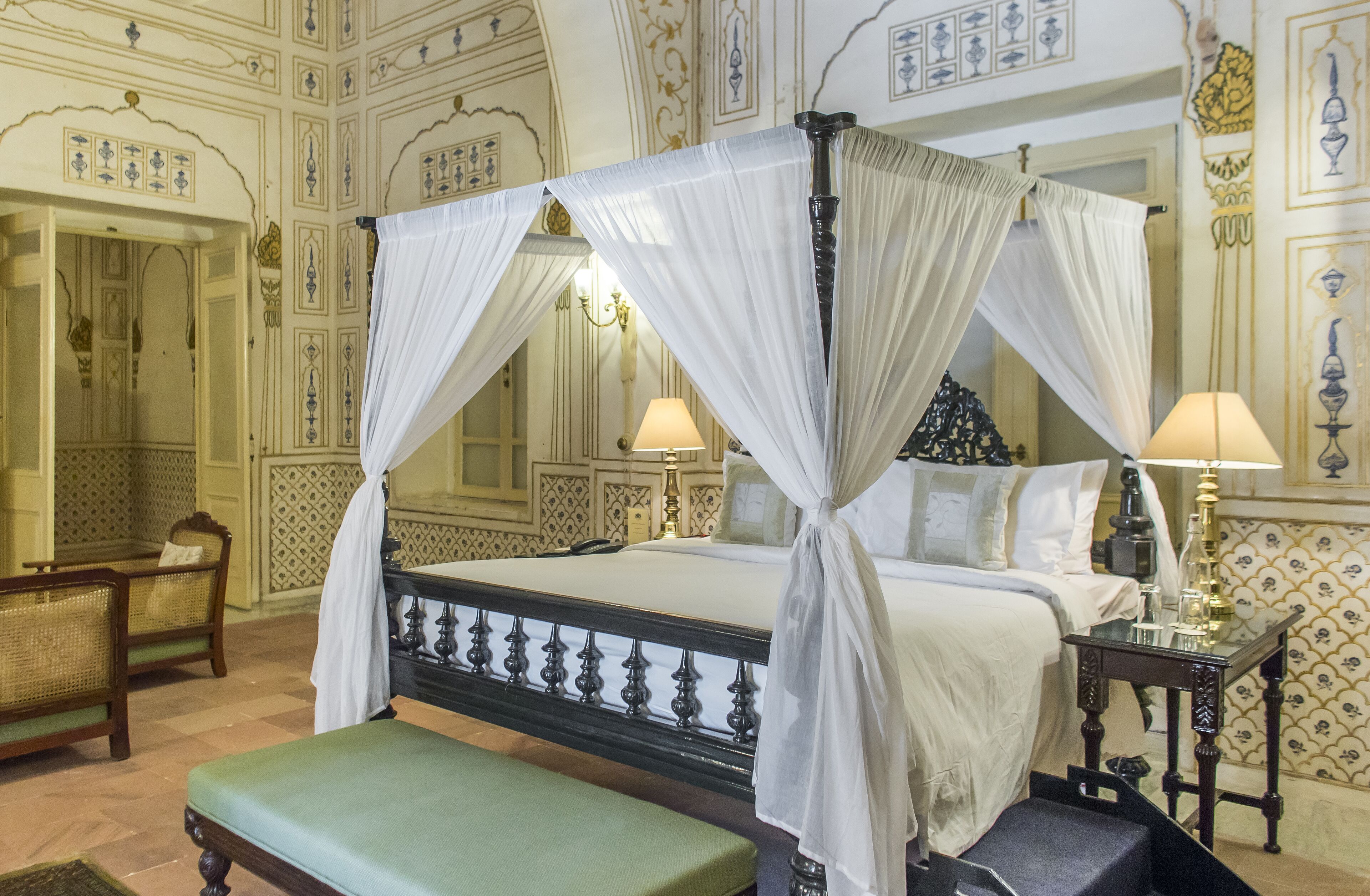 Una cama con dosel adornada con cortinas blancas, un banco verde, una mesita con lámpara y diseños ornamentales en las paredes.