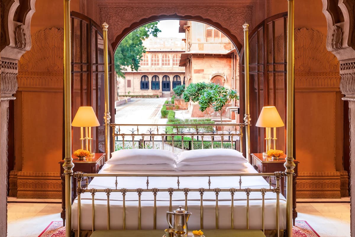Un lit recouvert d’une couverture verte, une table basse avec un service à thé, un tapis aux motifs rouges et noirs, et une vue sur une cour intérieure avec un bâtiment et de la verdure.