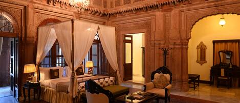 A luxurious bedroom with a four-poster bed, a sitting area with a sofa and armchair, and a chandelier.