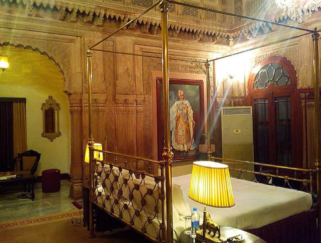 A four-poster bed with a lamp, a framed portrait on the wall, and a chandelier.