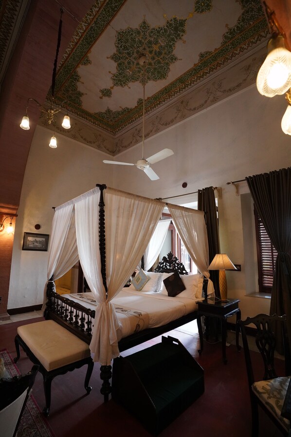 A four-poster canopy bed with a nightstand, a lamp, and a chair in a room with a ceiling fan and decorative ceiling.