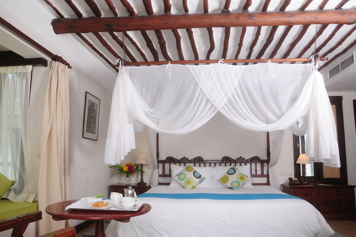 Quarto com cama sob um dossel, uma mesinha com itens de café da manhã e uma cadeira verde.