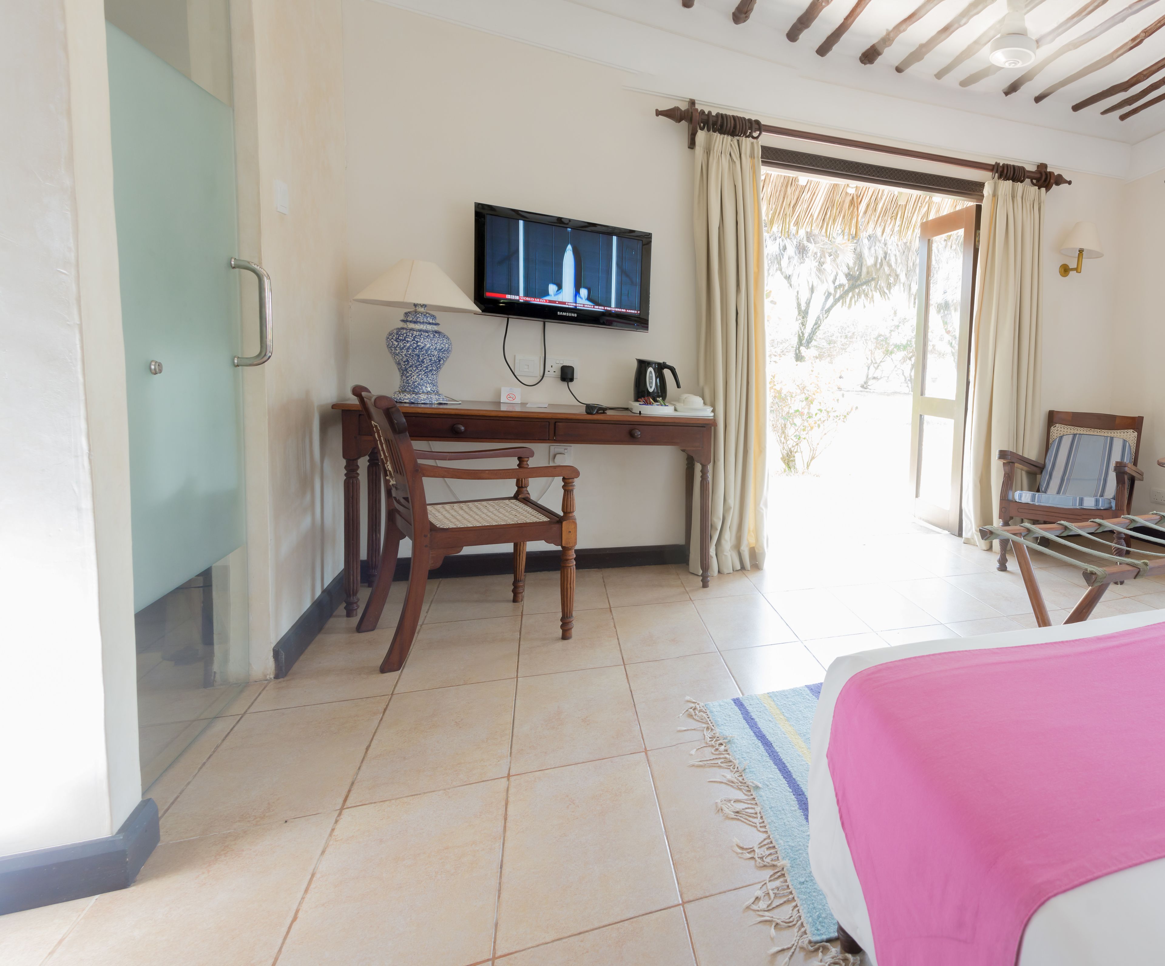 A hotel room with a wooden desk, a television, a chair, a bed with a pink blanket, and a view of the outdoors.