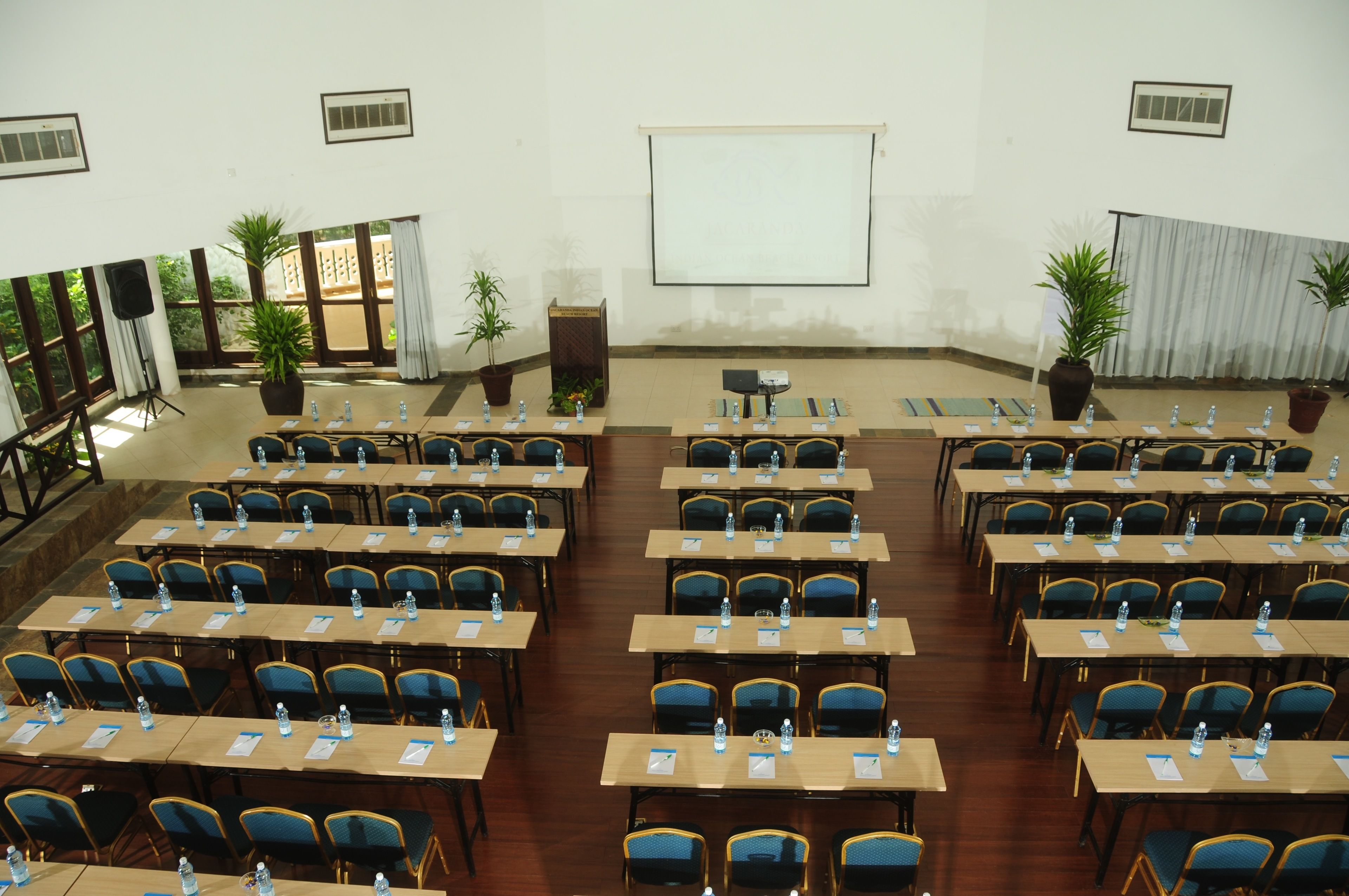 Une salle de conférence avec des rangées de tables et de chaises, un écran de projection et des plantes en pot.