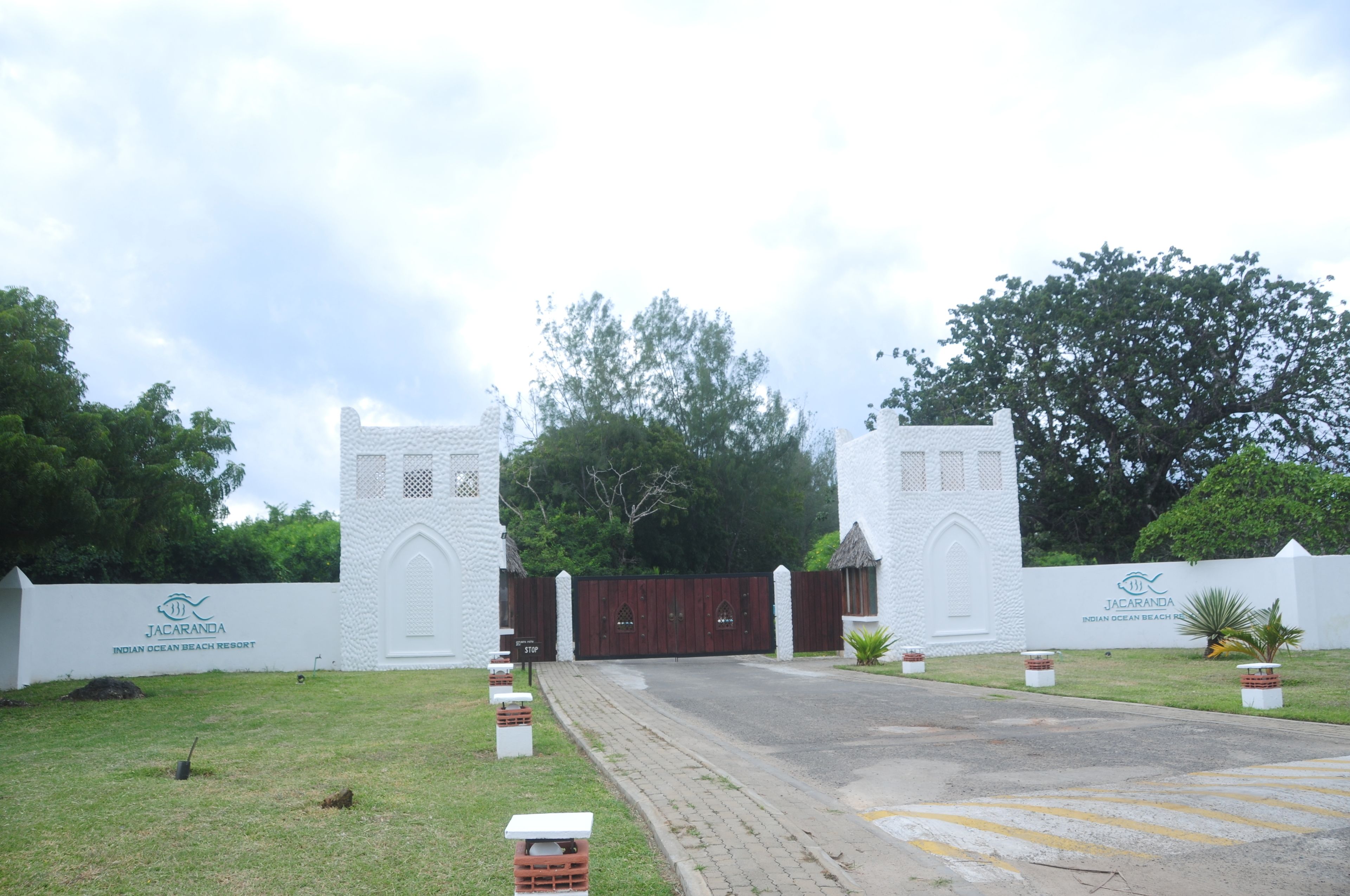 The entrance to Jacaranda Indian Ocean Beach Resort with a gated driveway, white walls, and greenery.