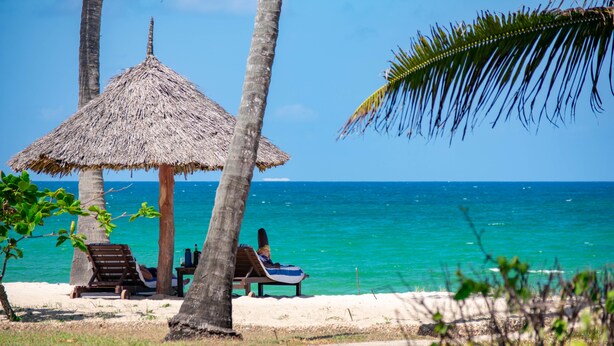 Escena de playa con una sombrilla de paja, dos sillas de madera y una mesita con una botella.