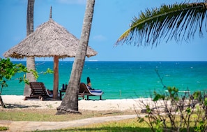 A beach scene with a thatched umbrella, two wooden lounge chairs, and a small table with a bottle. - Jacaranda Indian Ocean Beach Resort (Diani Beach)