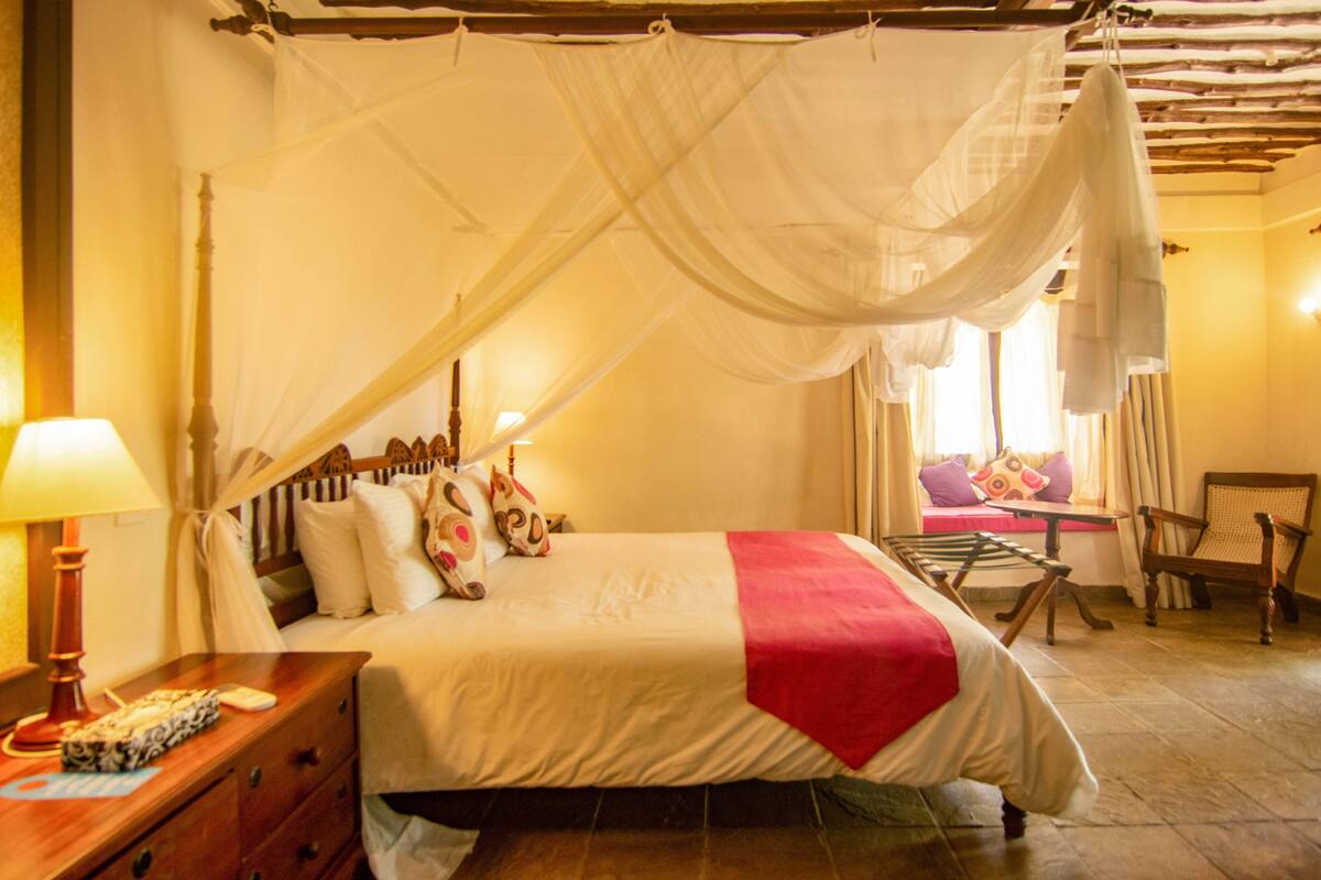 Quarto com cama de dossel, mesa de cabeceira, abajur e uma cadeira.