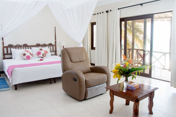 A hotel room with a bed, a reclining chair, a wooden table with a vase of flowers, and a view of palm trees through the window. - Jacaranda Indian Ocean Beach Resort (Diani Beach)