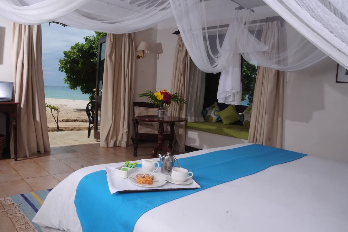 Quarto de hotel com cama, bandeja de café da manhã, vista para a praia e mesa com um vaso de flores.