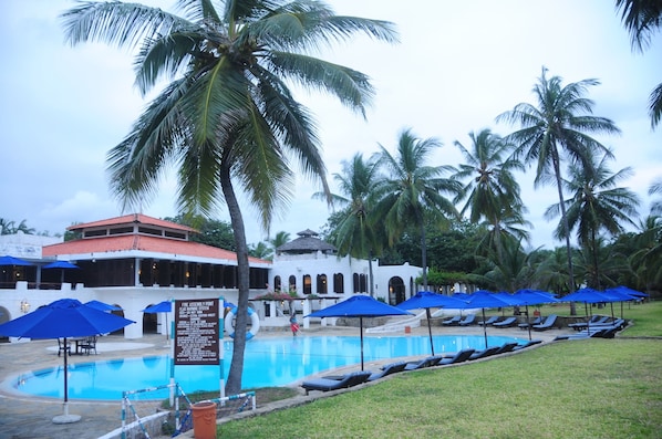 A pool area with lounge chairs and umbrellas, surrounded by palm trees and a building with a red roof. - Jacaranda Indian Ocean Beach Resort (Diani Beach)