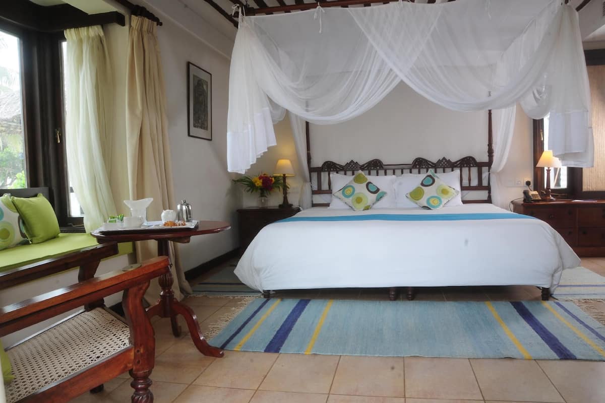 Quarto com cama, um banquinho, mesa de cabeceira, luminária e janela com cortinas.