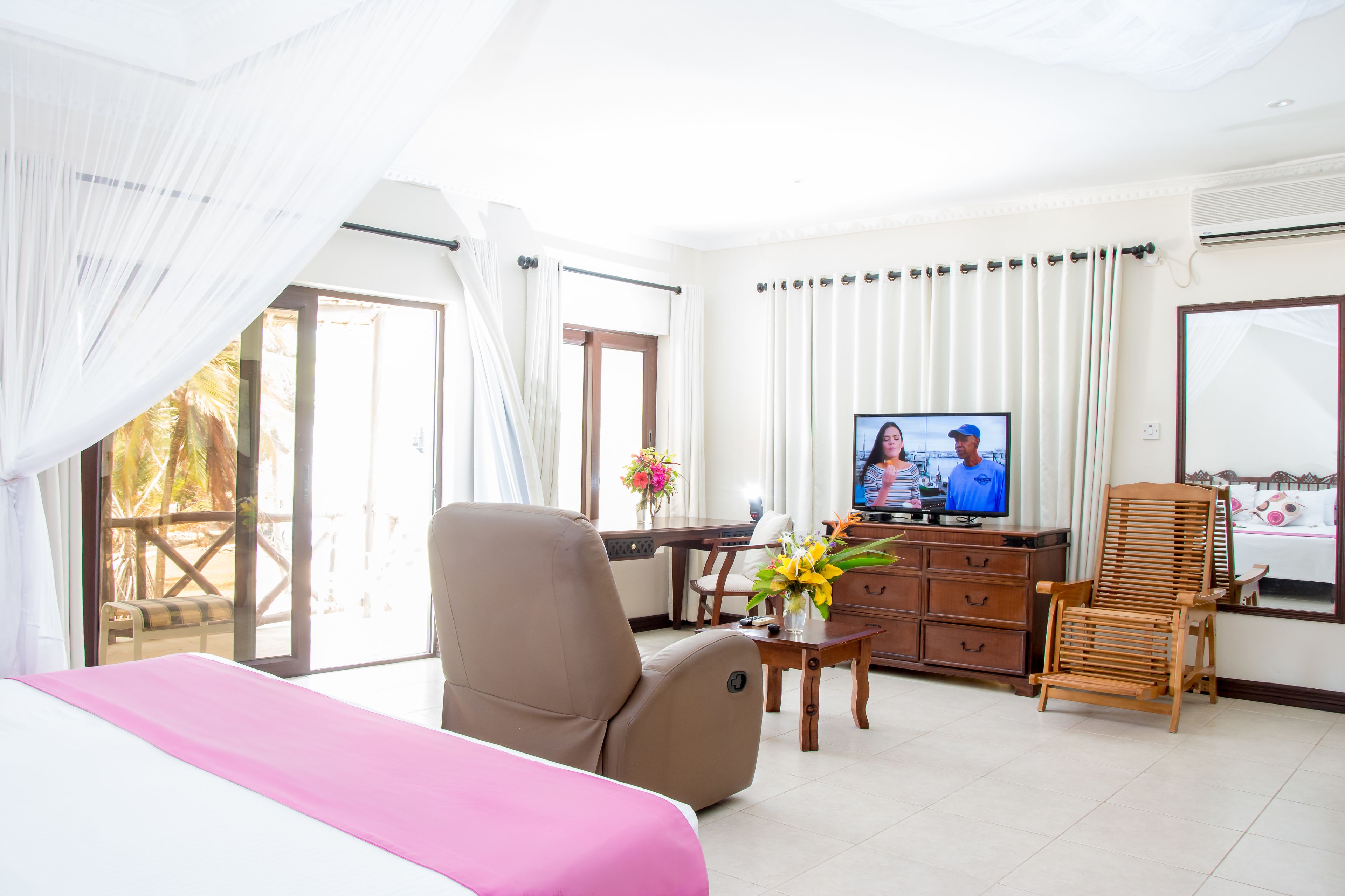 A hotel room with a bed, a wooden chair, a TV, a desk, and a window with curtains.