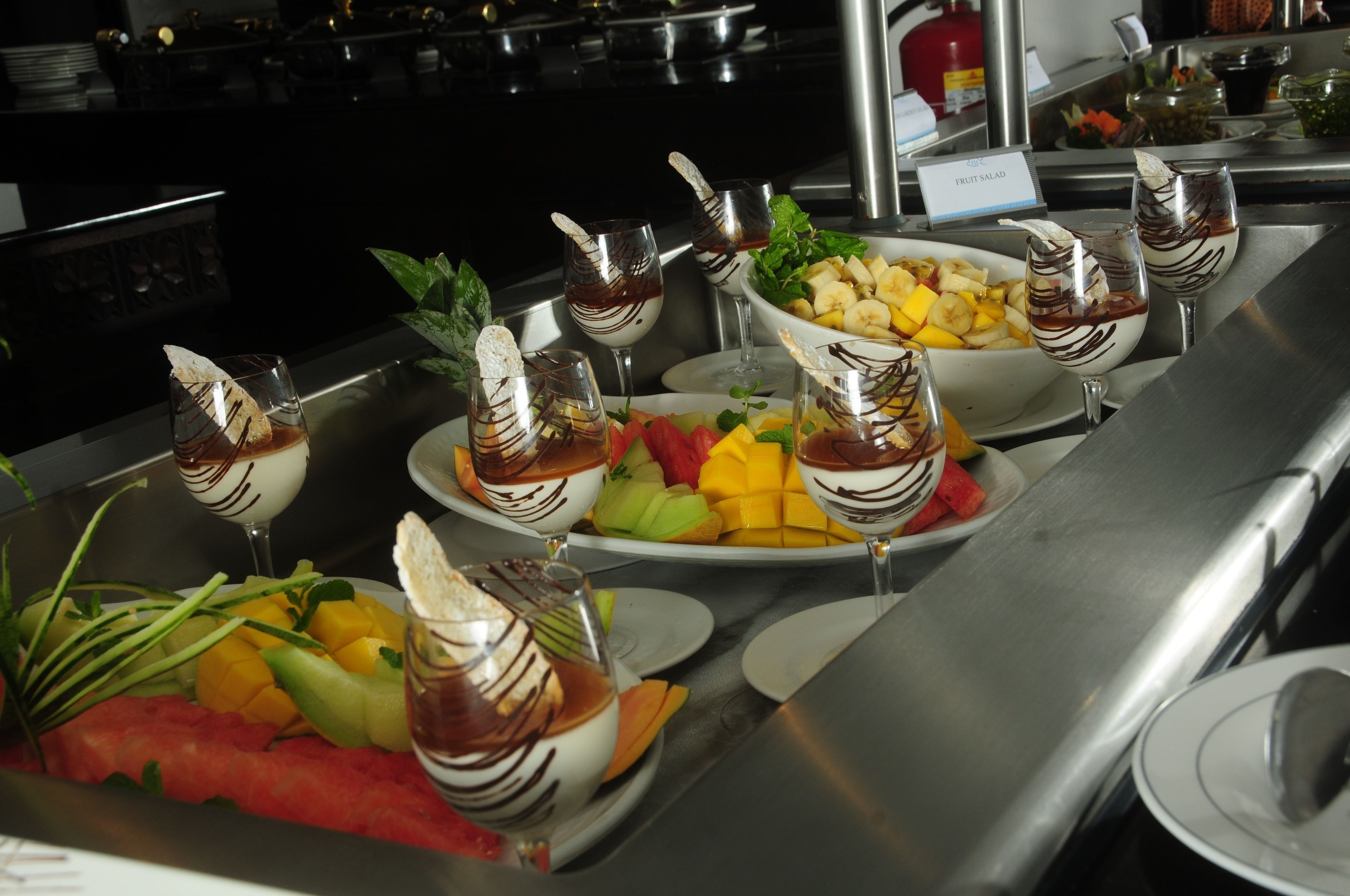 A buffet with various dishes and drinks, including fruit salad and a bowl of mixed fruits.