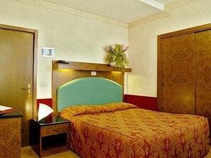 Habitación de hotel con cama, mesita de noche, armario de madera y estante con un jarrón de flores.