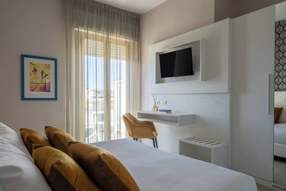 A modern hotel room with a large bed, a desk with a chair, a TV mounted on the wall, and a window with sheer curtains.