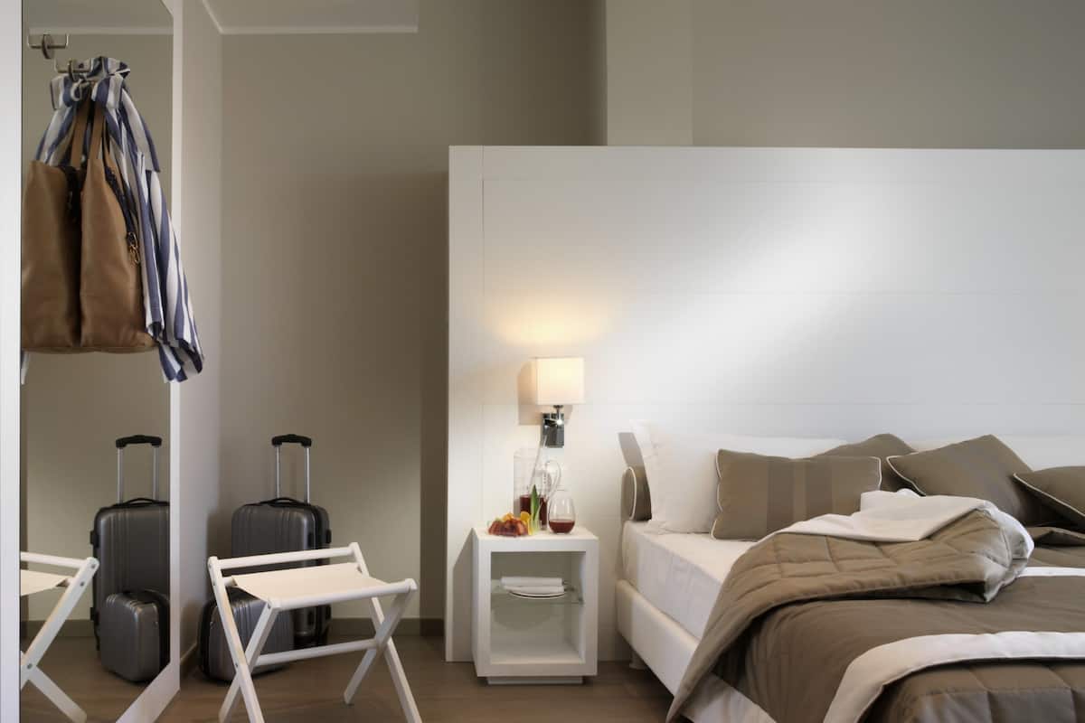 A modern hotel room with a bed, bedside table, lamp, and a seating area with a suitcase and a bag.