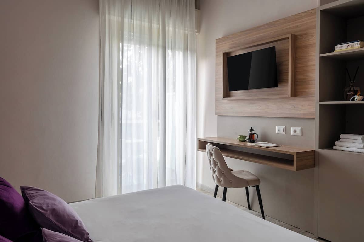 A modern bedroom with a bed, a desk with a chair, a TV mounted on a wooden shelf, and a window with sheer curtains.