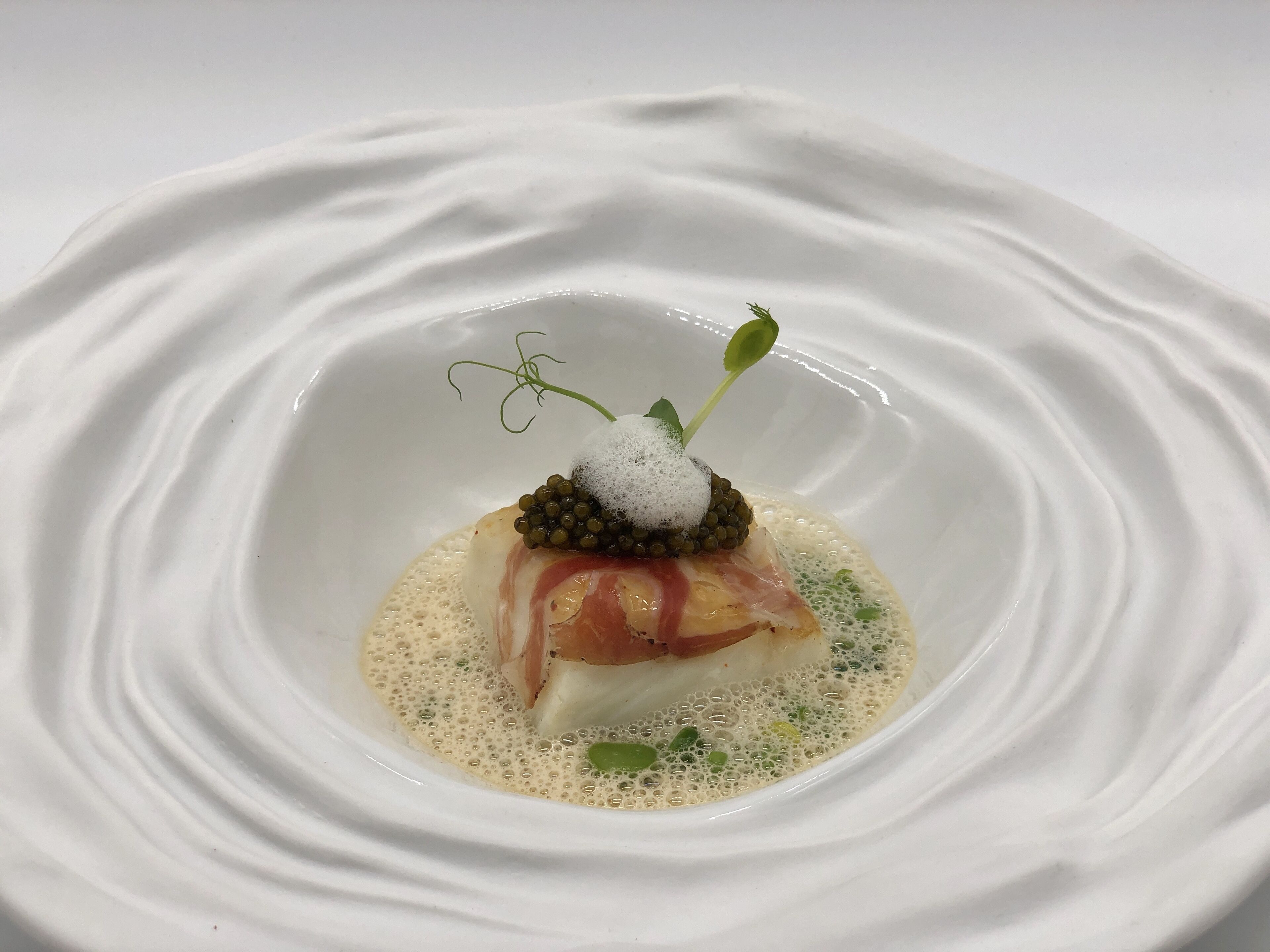 Ein weißer Teller mit einem zentral platzierten Gericht, bestehend aus Fisch, Kaviar und einem Zweig von grünem Kräutern.
