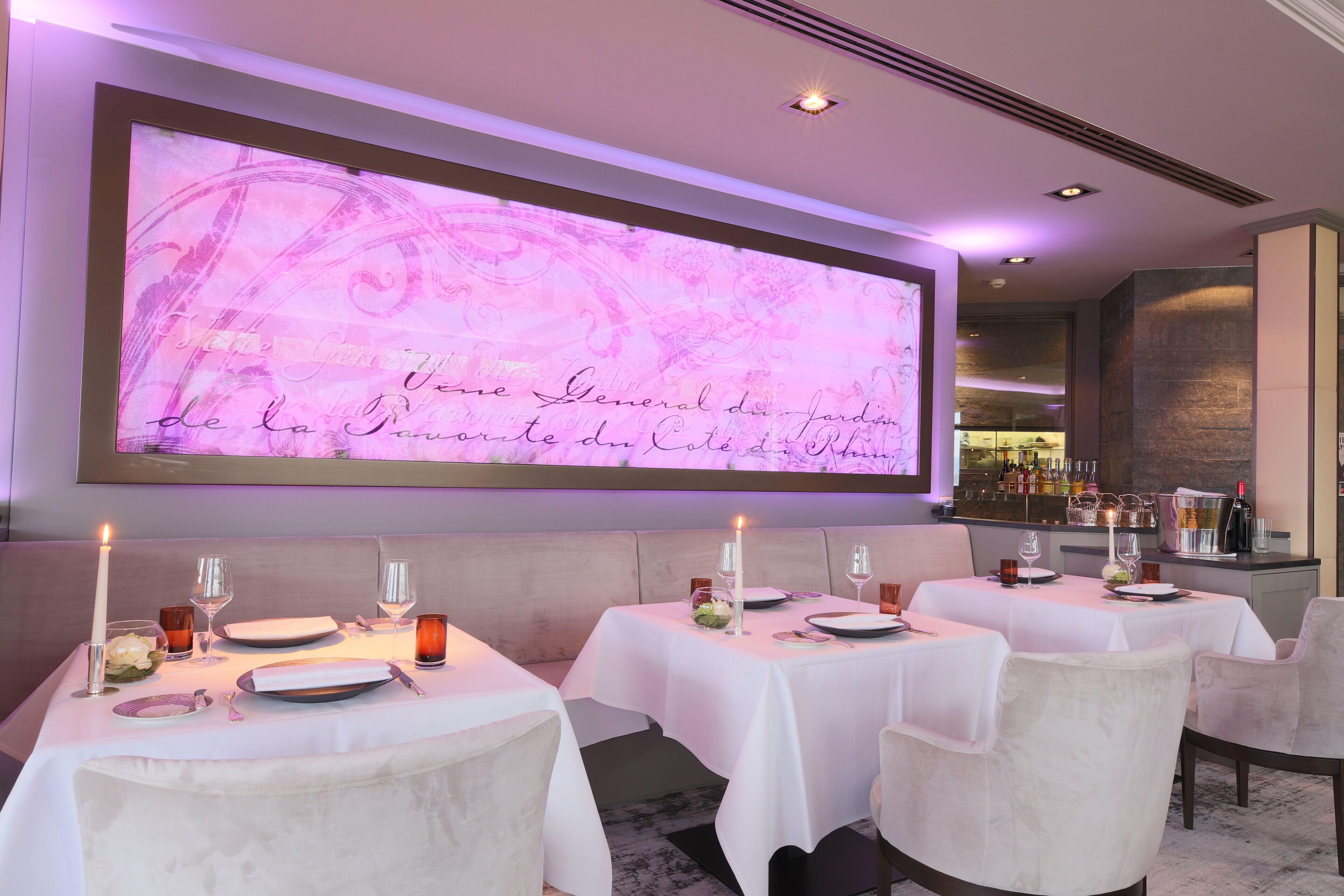 A dining area with tables set for two, featuring white tablecloths and candles. A large illuminated sign with cursive writing is visible on the wall.