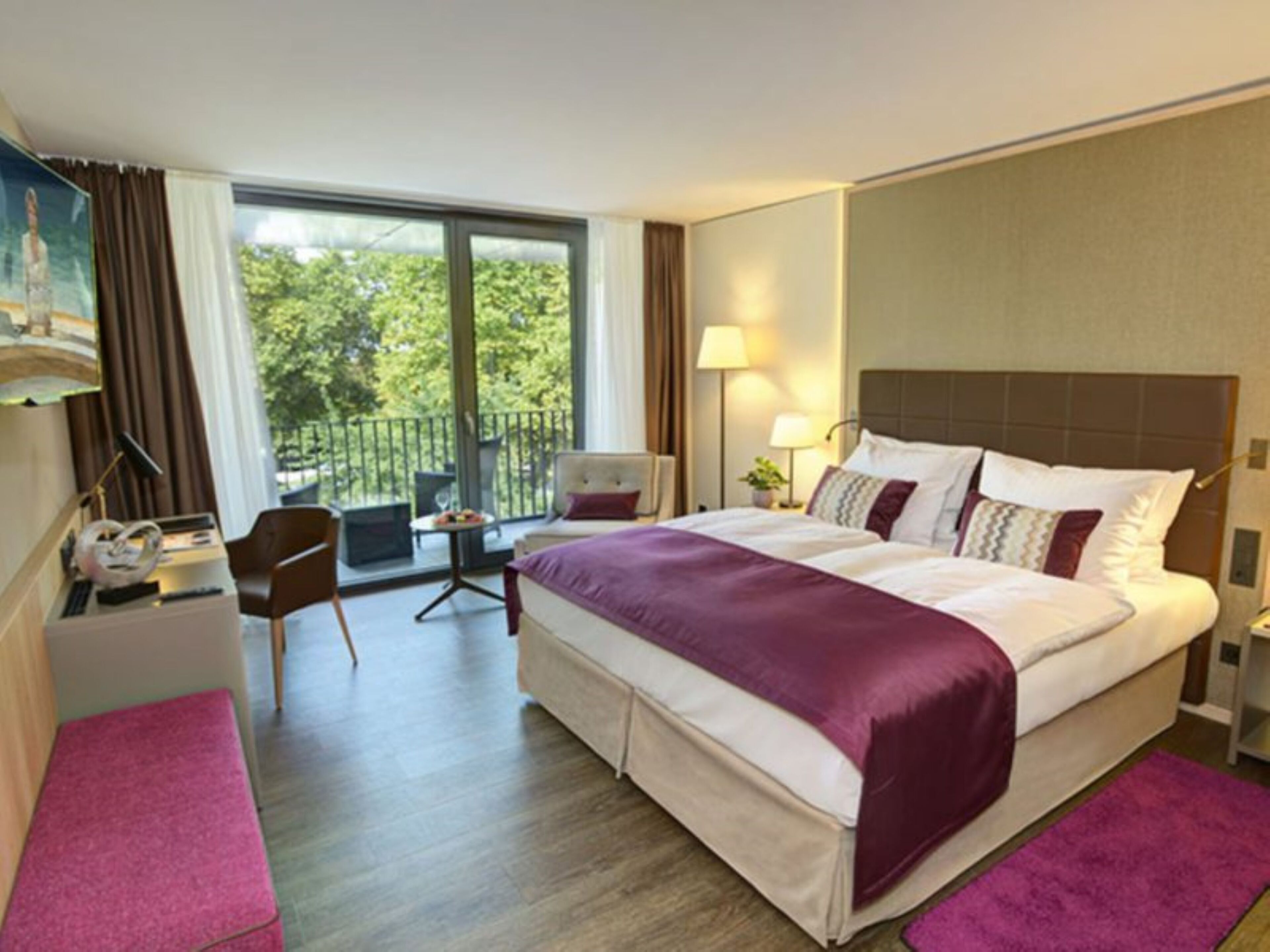 Habitación de hotel con una cama grande, un escritorio, una silla y un balcón con vistas a árboles.