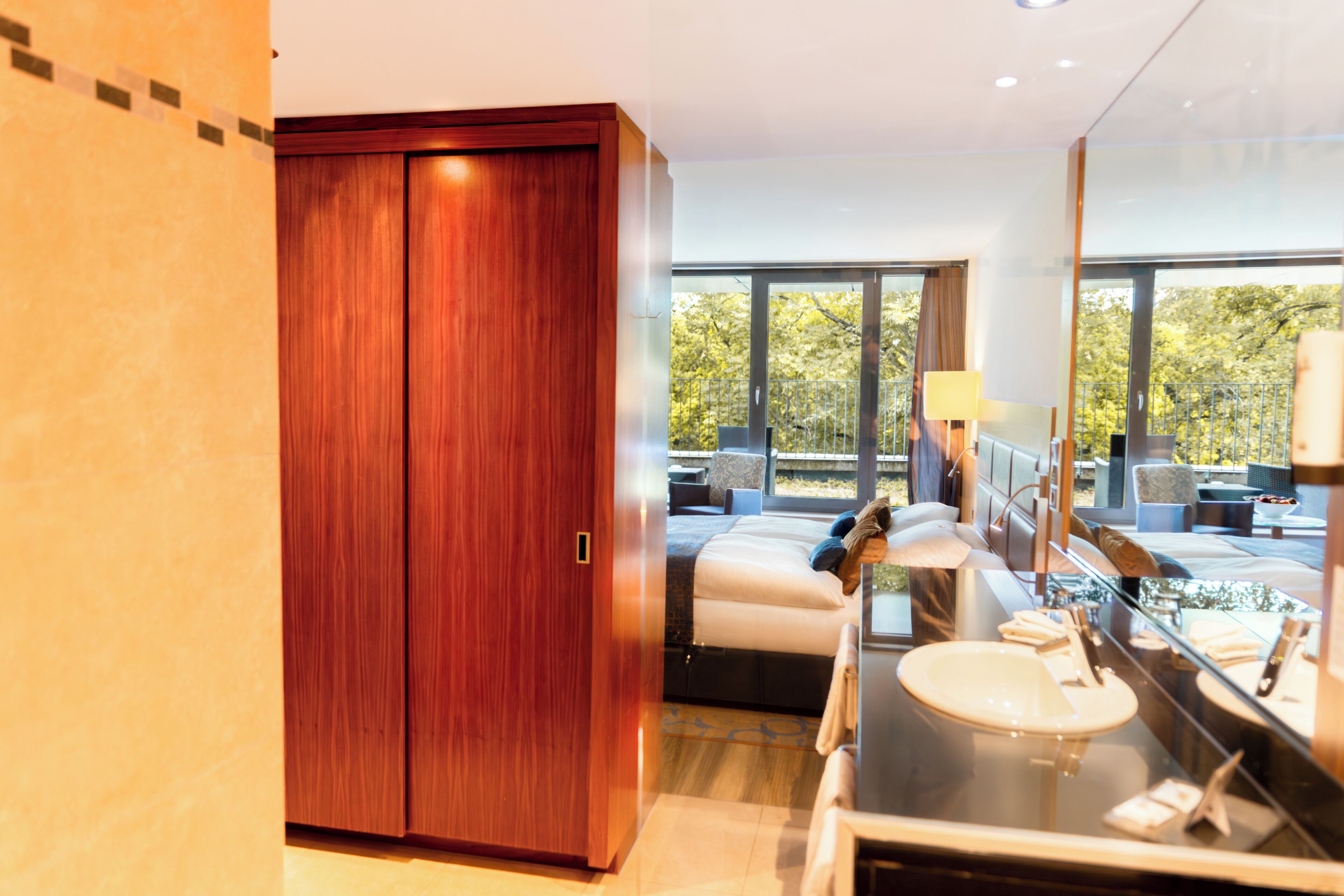 Habitación de hotel moderna con un amplio armario de madera, cama con vistas al exterior, baño con un espejo grande y una zona de estar con mesa y sillas.