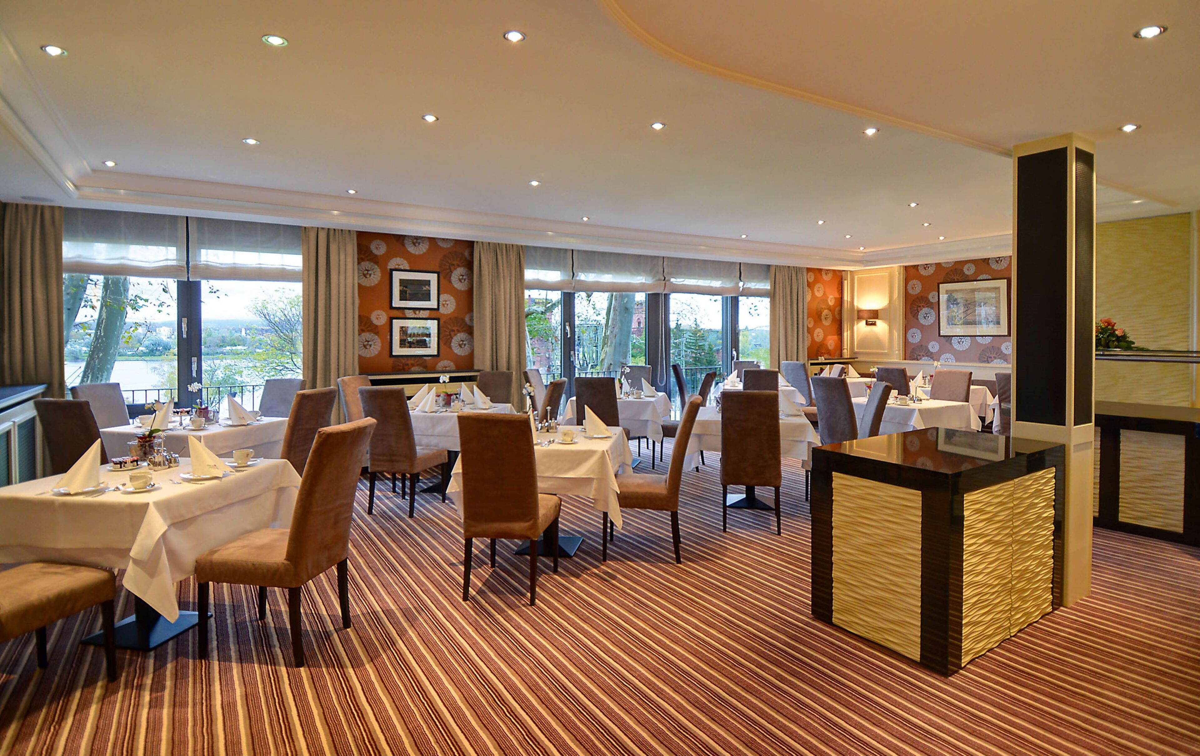 A hotel dining area with tables set for guests, large windows offering a view, and a central bar area.