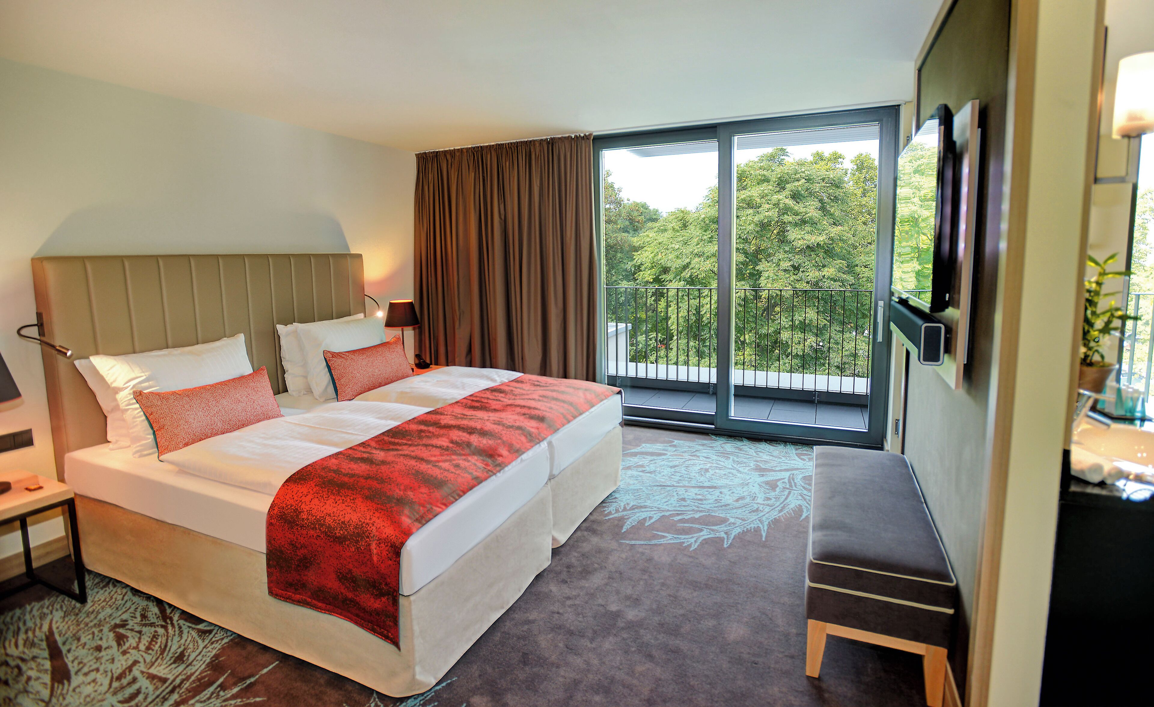 A hotel room with a large bed, a bench, a TV, and a balcony with a view of trees.