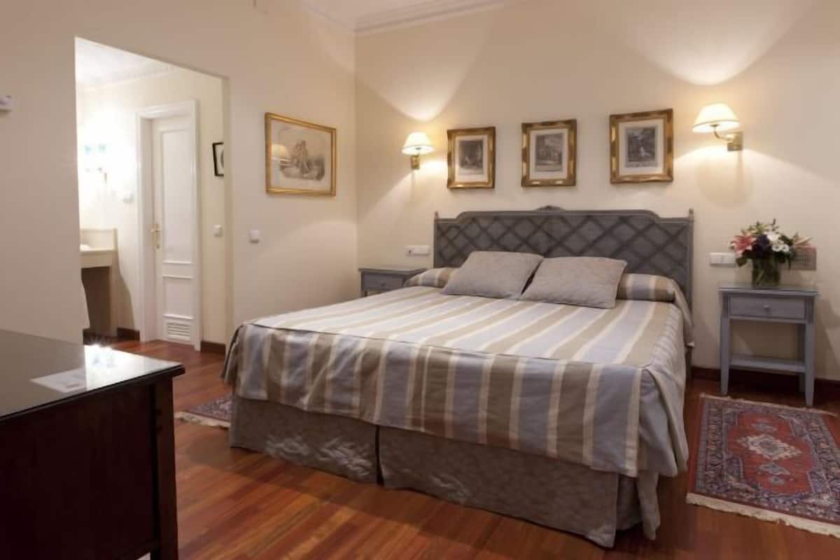 A bedroom with a bed, bedside tables, wall-mounted lamps, framed pictures, and a rug.