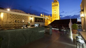 An outdoor bar area with a view of a tall, illuminated tower in the background. - Hotel Doña Maria (Seville)