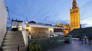 A rooftop bar with a view of a historic tower, outdoor seating, and a staircase leading to the bar area. - Hotel Doña Maria (Seville)