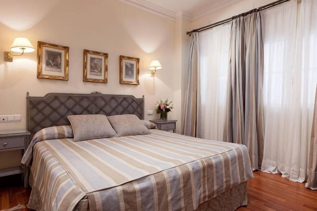 A bedroom with a bed, bedside tables, wall-mounted lamps, framed pictures, and a window with curtains.