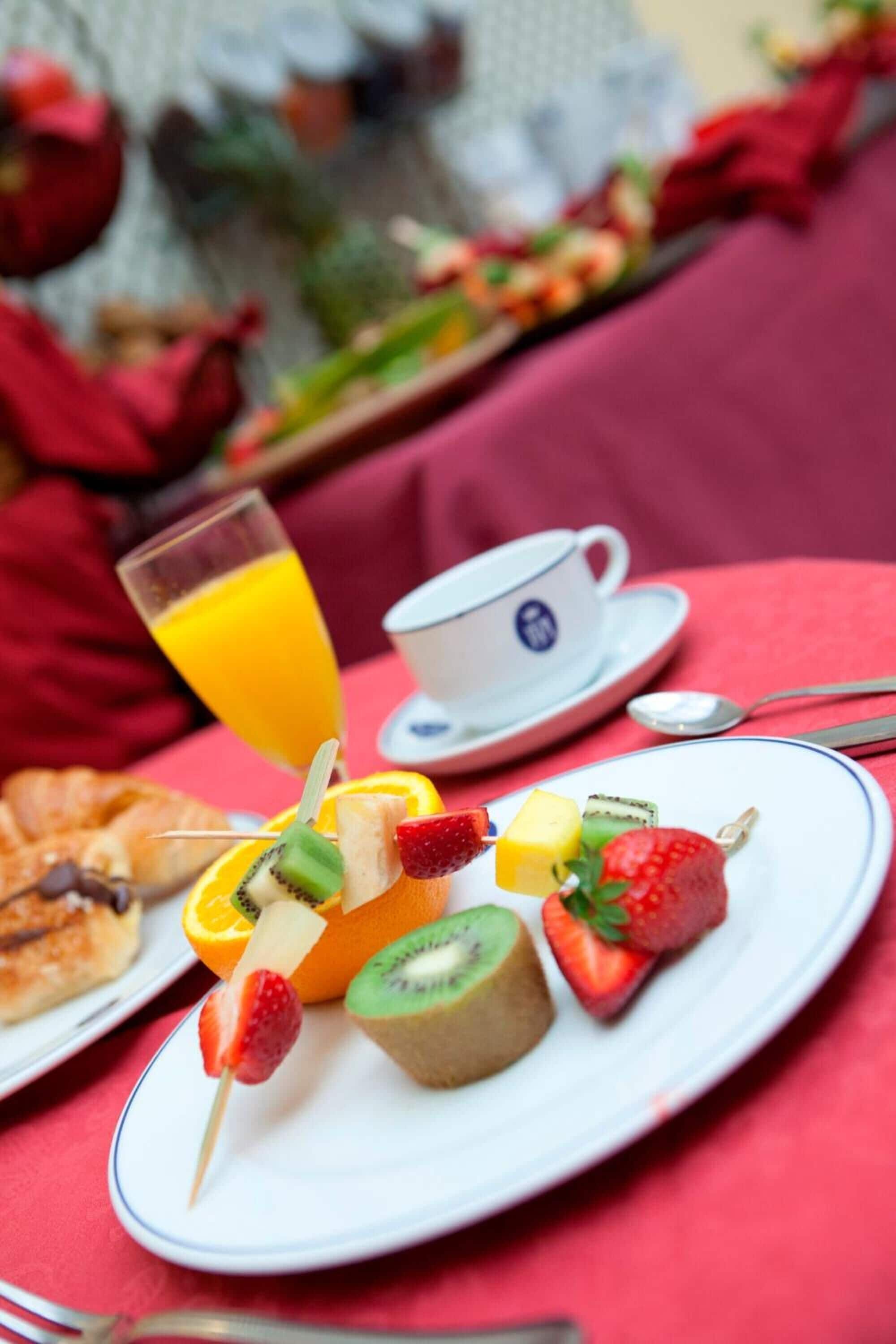 Une table mise pour le petit-déjeuner avec des brochettes de fruits, un verre de jus d’orange et une tasse de thé, le tout posé sur une nappe rouge.