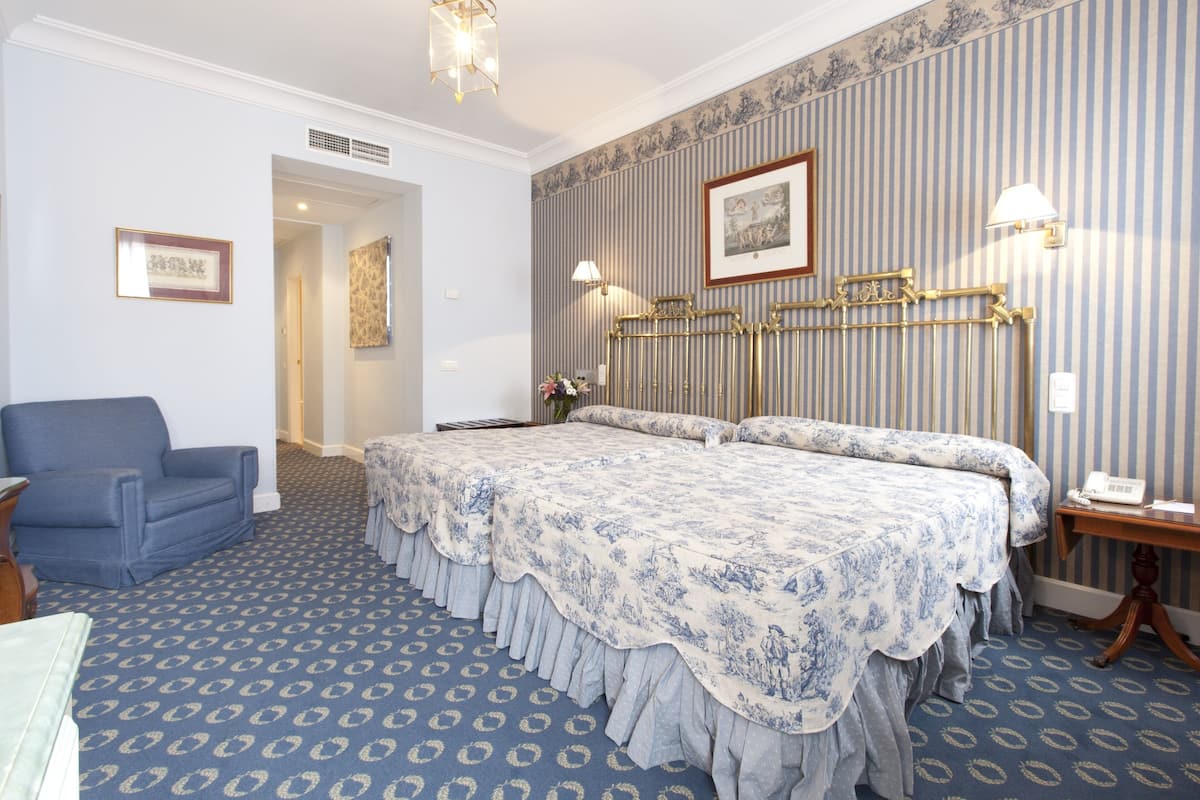 A hotel room with a large bed, a blue armchair, a small table with a lamp, and a framed picture on the wall.