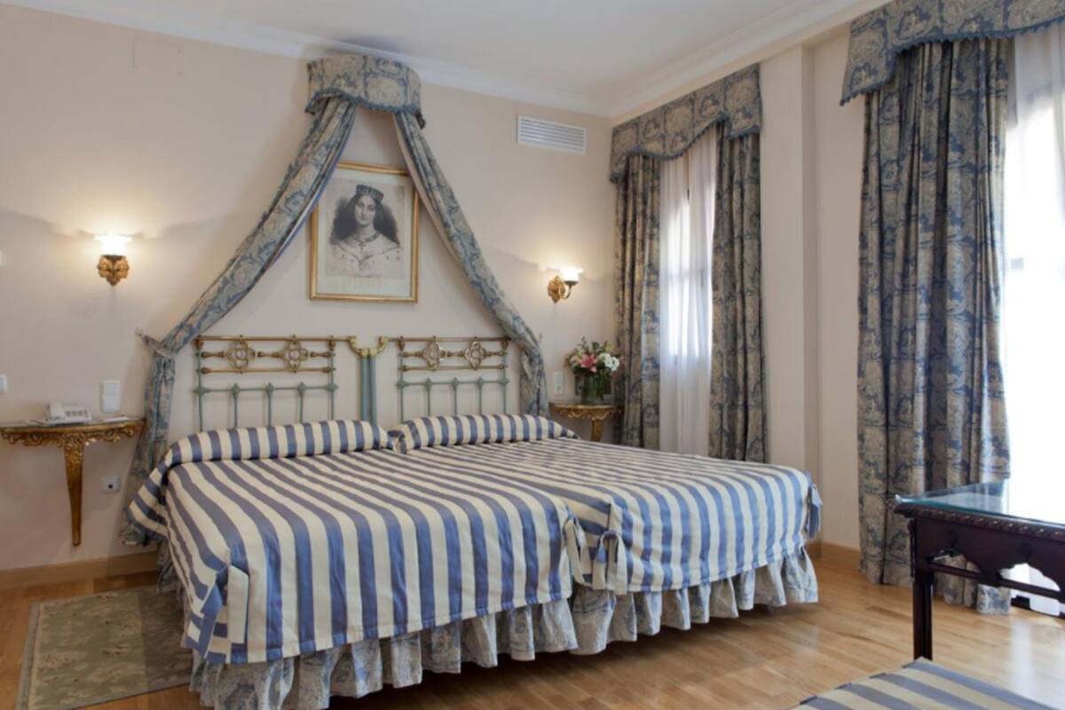 A bedroom with a large bed, a headboard with a decorative design, two wall-mounted lamps, a framed picture above the bed, and a window with curtains.