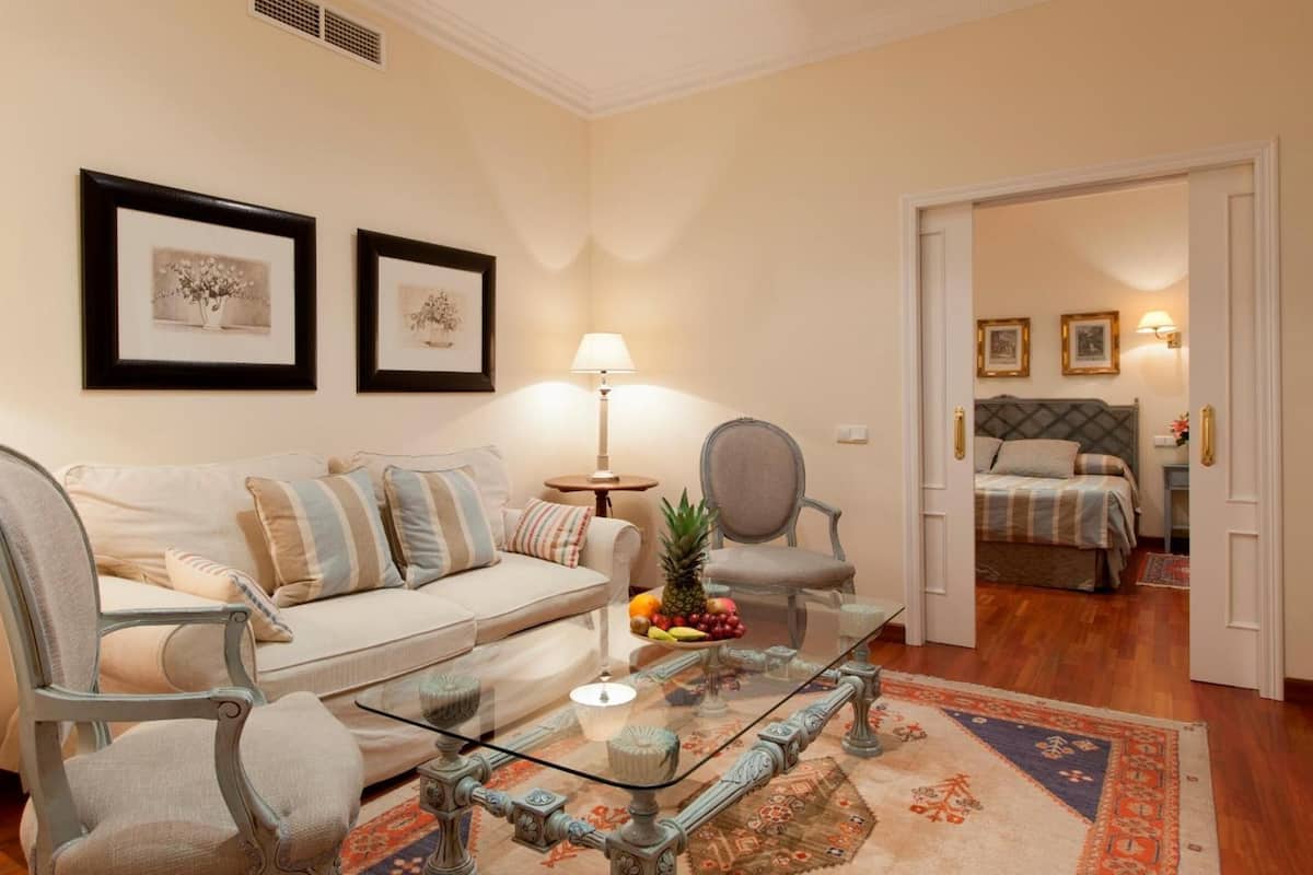 A living room with a sofa, armchairs, a glass coffee table, and a dining table. There are framed pictures on the wall and a lamp on a side table.