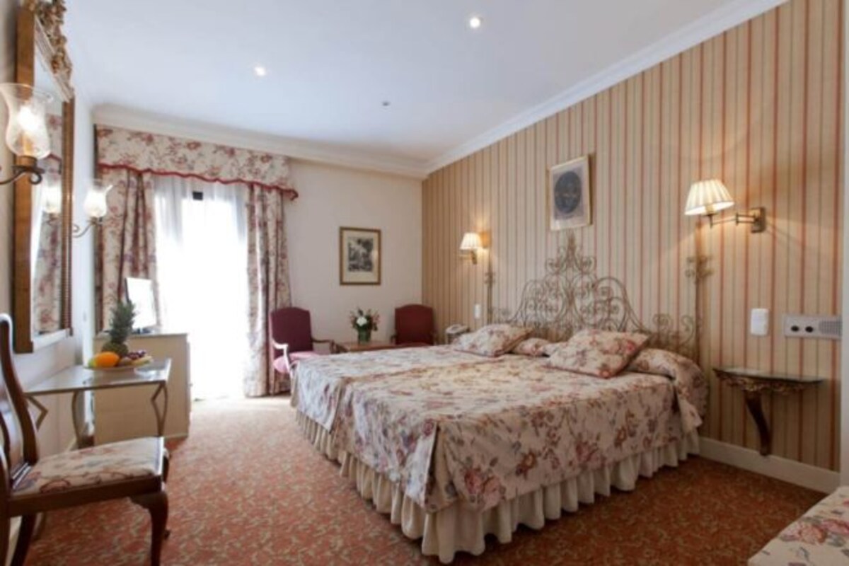A hotel room with a double bed, a desk with a chair, a small table, and a mirror.