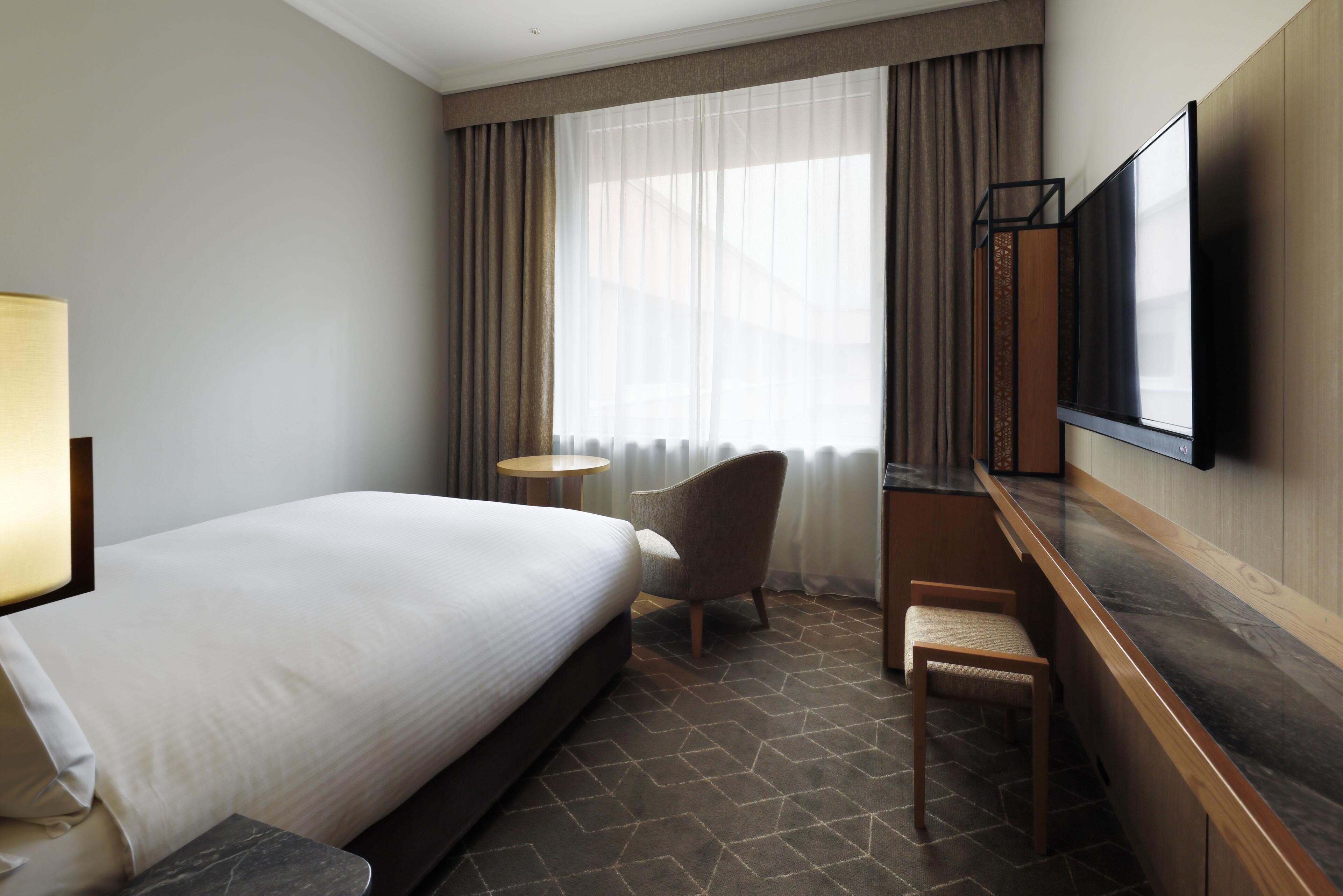 A hotel room with a large bed, a desk, a chair, a TV, and a window with curtains.