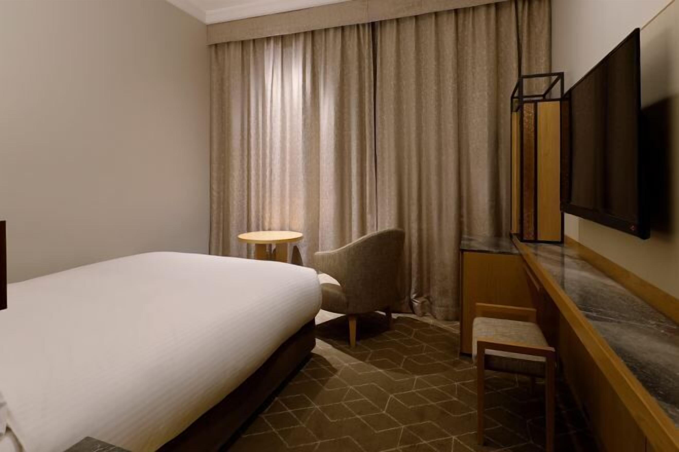 A hotel room with a bed, a chair, a small table, a TV, and a window with curtains.