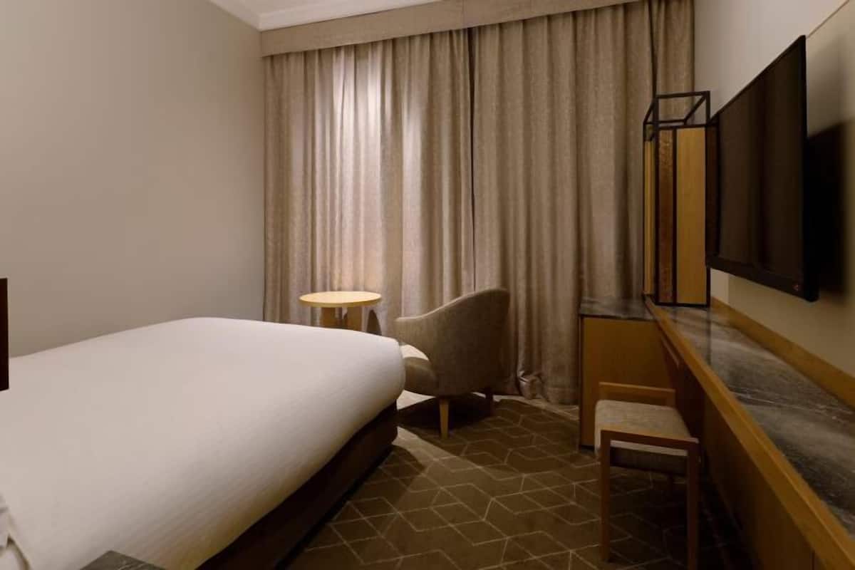 Une chambre d’hôtel avec un lit, une chaise, une petite table, un téléviseur et une fenêtre avec des rideaux.