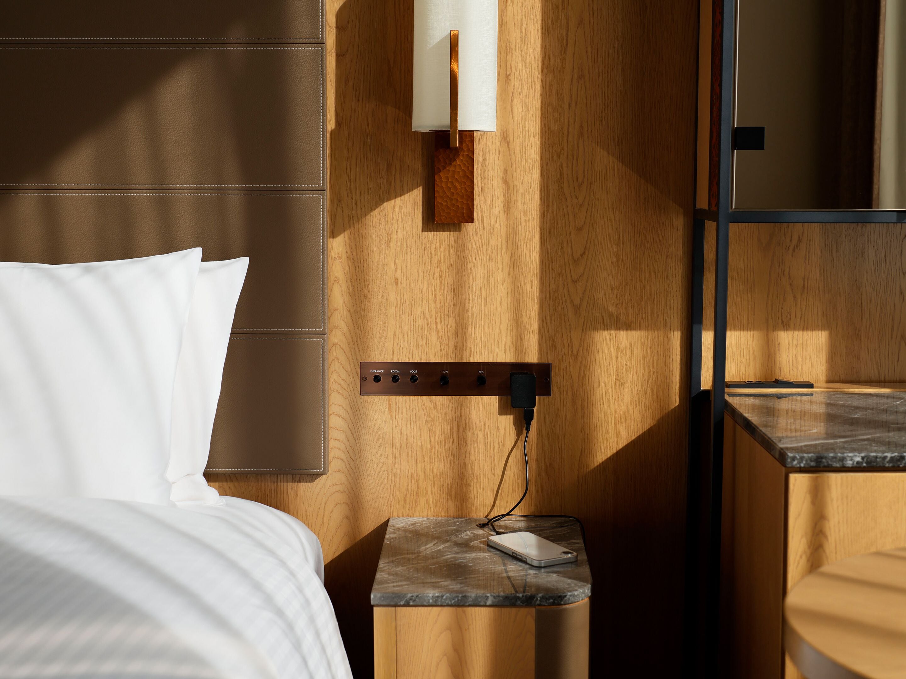 A hotel room with a bed, a bedside table with a phone charger, a wall-mounted light, and a mirror.