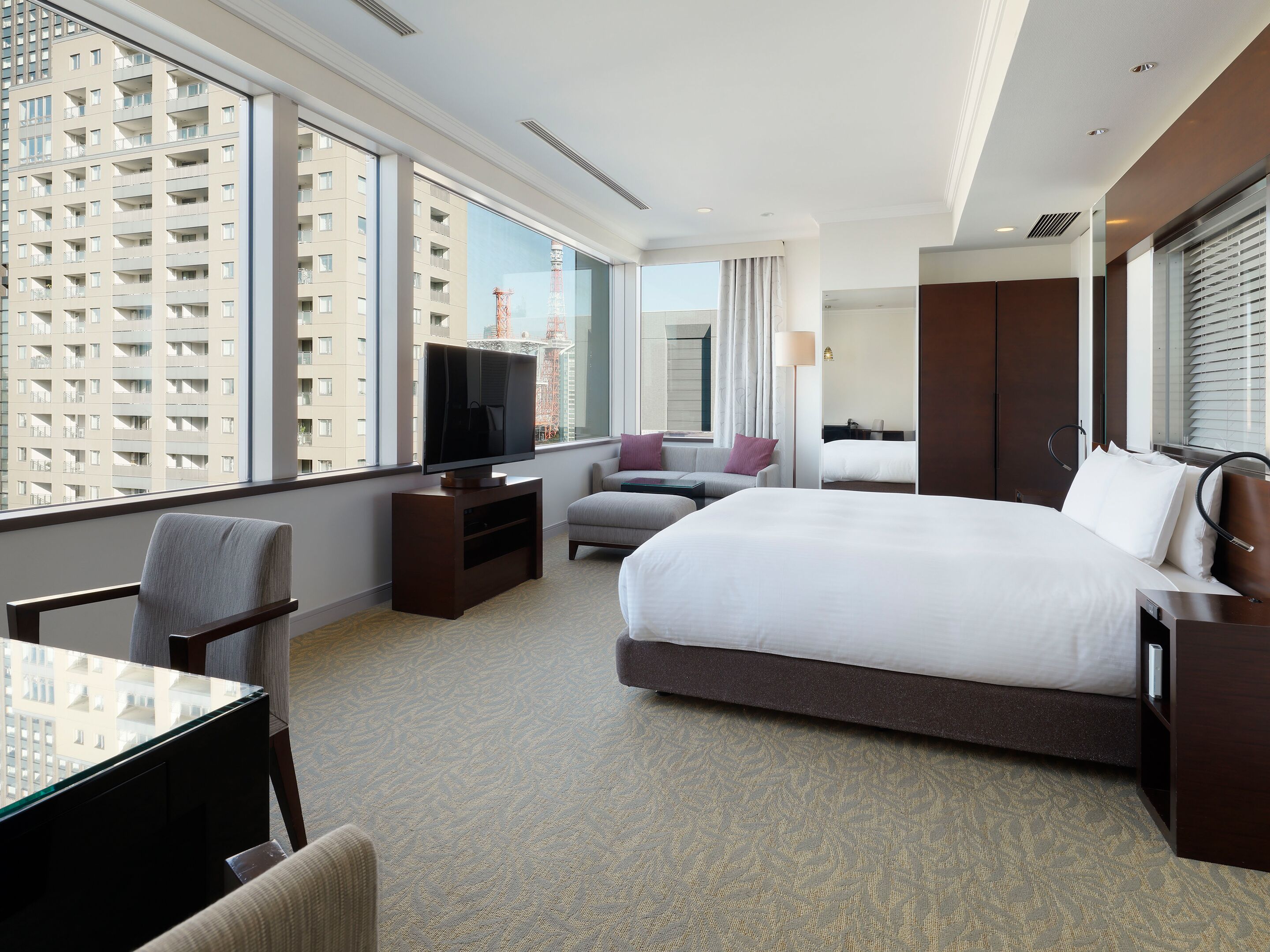 A hotel room with a large bed, a television, a desk, and a view of high-rise buildings.