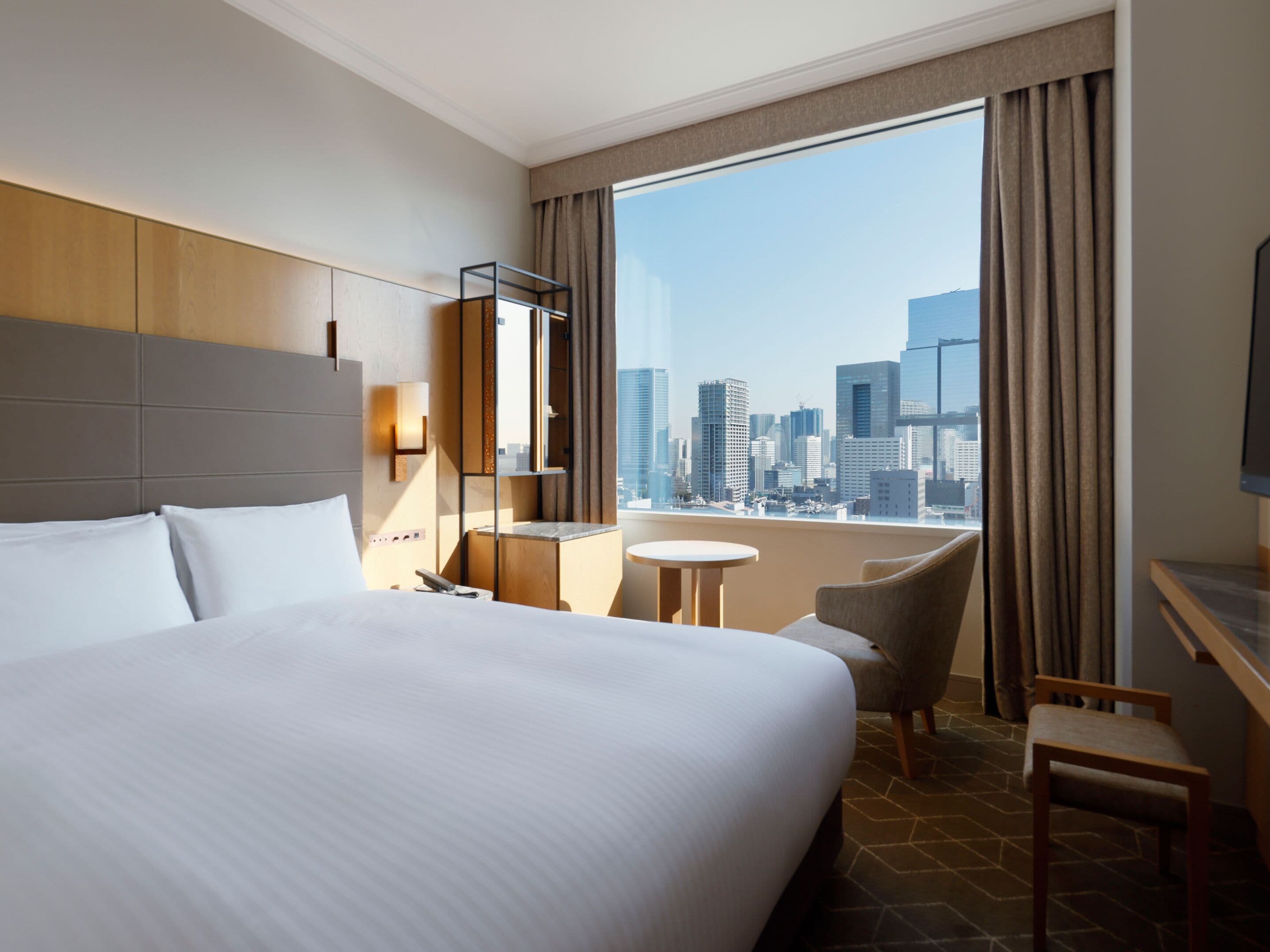 Ein Hotelzimmer mit einem großen Bett, einem Schreibtisch, einem Sessel, einem kleinen Tisch und Blick auf die Stadt durch ein großes Fenster.