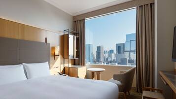 Ein Hotelzimmer mit einem großen Bett, einem Schreibtisch, einem Sessel, einem kleinen Tisch und Blick auf die Stadt durch ein großes Fenster.