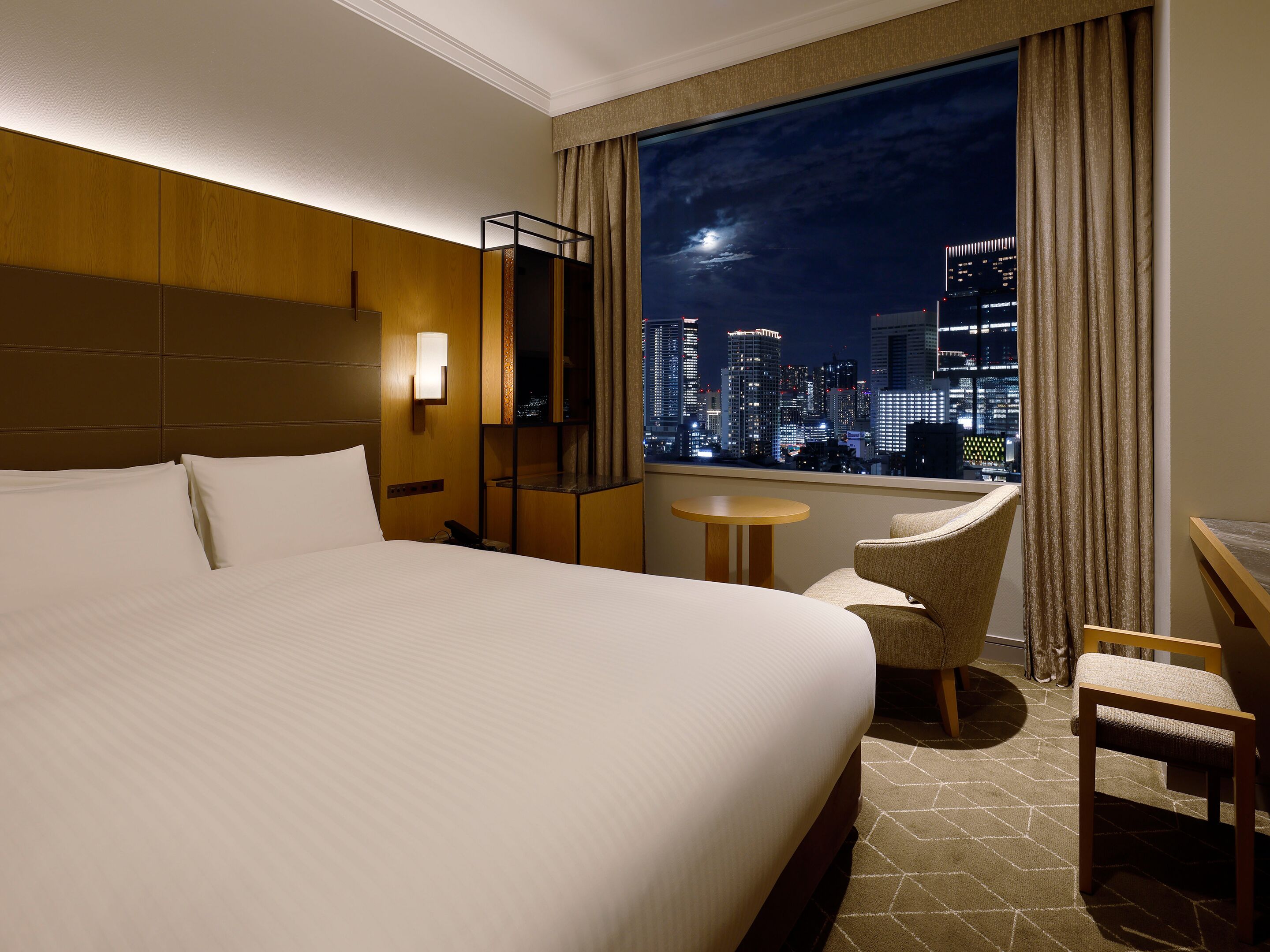 A hotel room with a large bed, a small table, a chair, a nightstand, and a window with a city view.