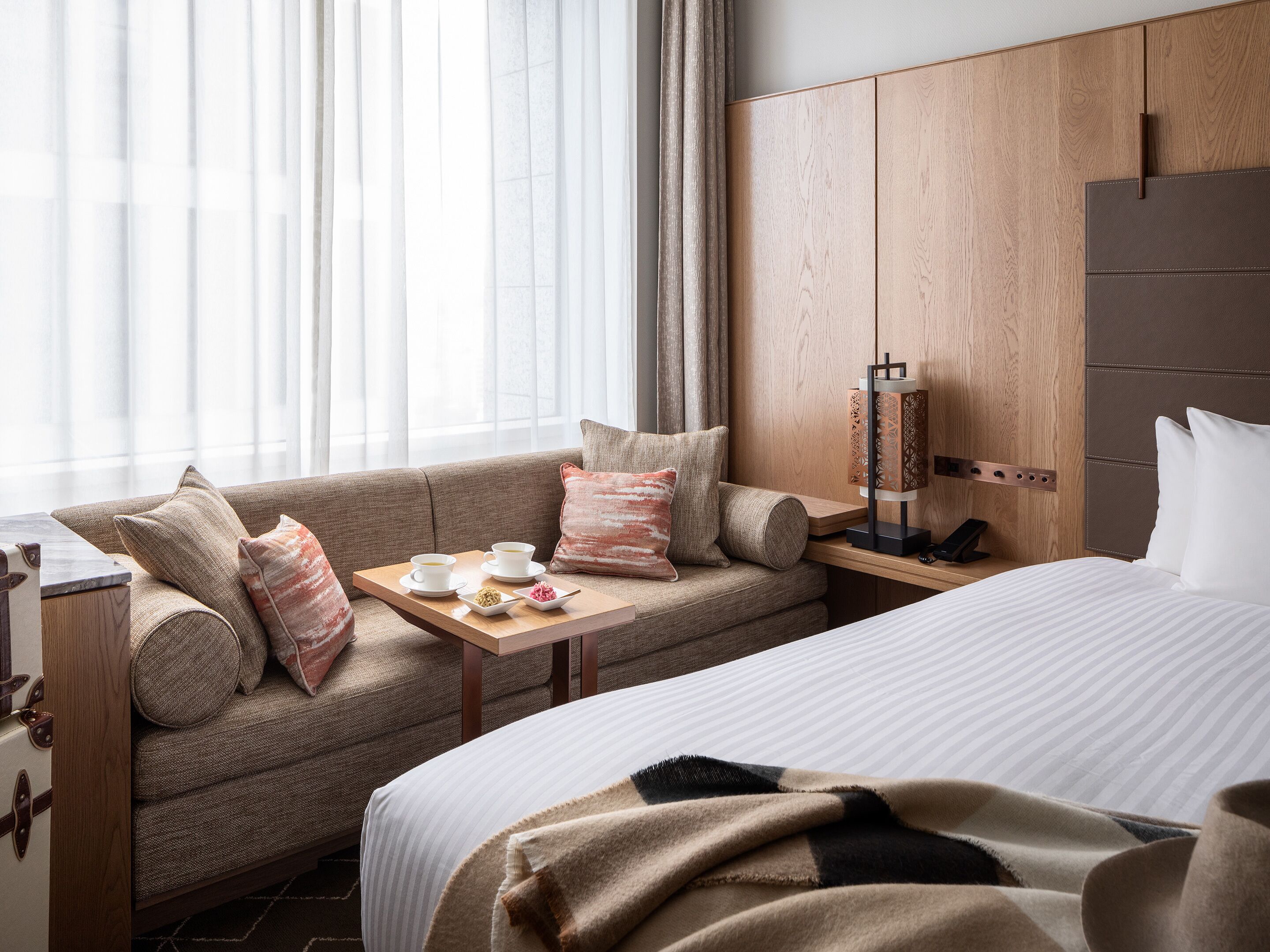 A hotel room with a sofa, a bedside table with a tray of tea and snacks, a lamp, and a bed with a striped duvet.