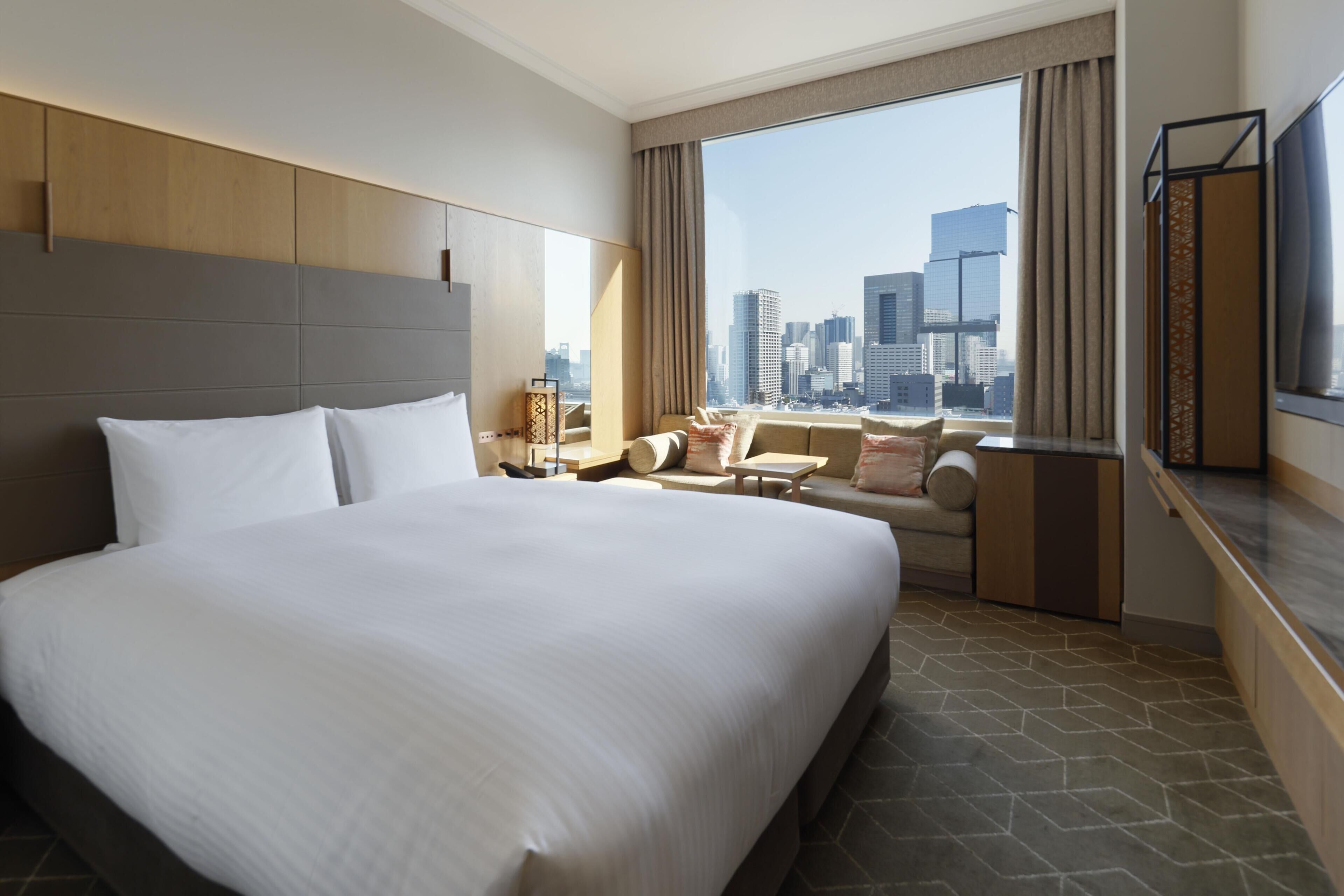 Habitación de hotel con una cama grande, un sofá, una mesita y vistas a un horizonte urbano a través de la ventana.