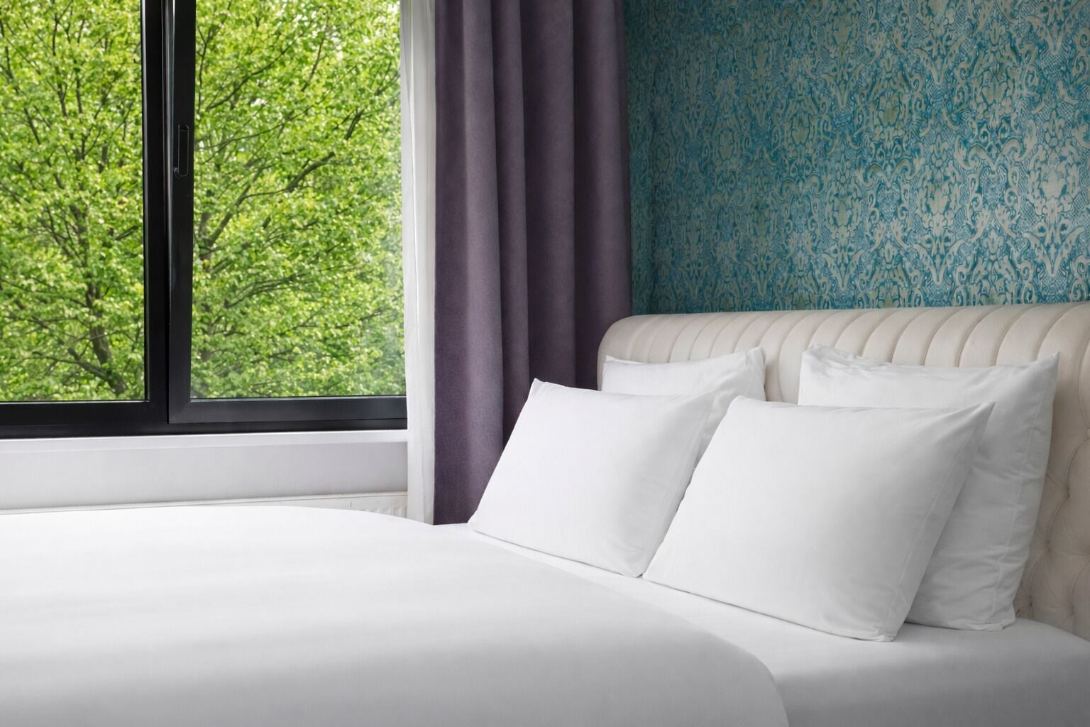 A bed with white linens, a window with a view of trees, and a patterned wall.