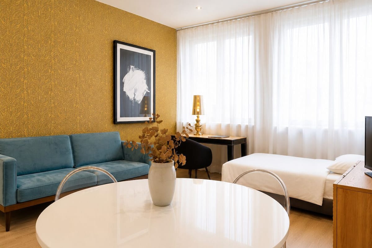 Hotelkamer met een blauwe bank, een rond tafeltje met een vaas, een bed, een bureau met een lamp en een televisie.